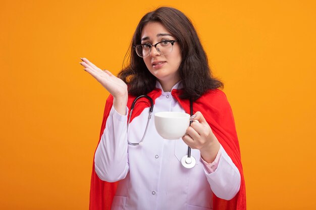 Beeindruckte junge Superheldin in Arztuniform und Stethoskop mit Brille, die eine Tasse Tee hält und nach vorne schaut, die leere Hand isoliert an der Wand mit Kopierraum zeigt