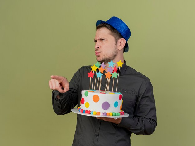 beeindruckte jungen Party-Typ, der schwarzes Hemd und blauen Hut hält, der Kuchen und Punkte an der Seite lokalisiert auf olivgrün hält