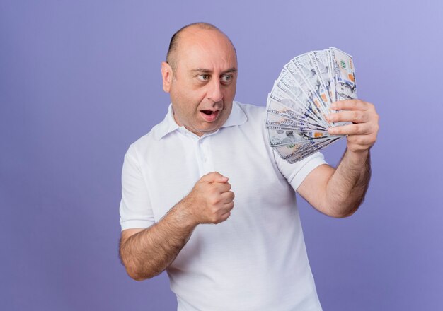 Beeindruckte lässige reife Geschäftsmann halten und betrachten Geld und geballte Faust lokalisiert auf lila Hintergrund mit Kopienraum