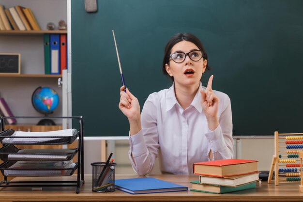 beeindruckte Punkte auf junge Lehrerin, die eine Brille trägt und einen Zeiger hält, der mit Schulwerkzeugen im Klassenzimmer am Schreibtisch sitzt