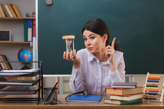 beeindruckte Punkte auf junge Lehrerin mit Sanduhr, die mit Schulwerkzeugen im Klassenzimmer am Schreibtisch sitzt