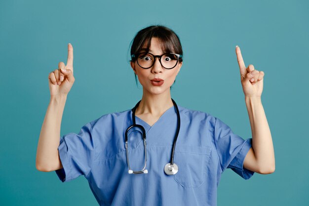 Beeindruckte Punkte auf junge Ärztin mit einheitlichem Fith-Stethoskop isoliert auf blauem Hintergrund