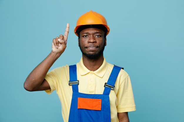 Beeindruckte Punkte auf jungen afroamerikanischen Baumeister in Uniform isoliert auf blauem Hintergrund