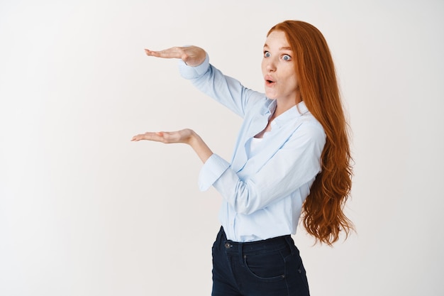Beeindruckte rothaarige Frau, die ein großes Objekt mit Händen zeigt, etwas Großes misst und überrascht nach links schaut, weiße Wand