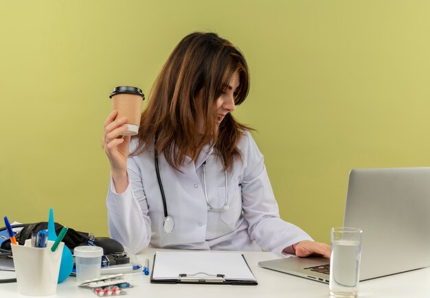 Beeindruckte Ärztin mittleren Alters, die medizinische Robe und Stethoskop trägt, die am Schreibtisch mit medizinischen Werkzeugen und Zwischenablage unter Verwendung des Laptop hält, der Plastikkaffeetasse isoliert hält