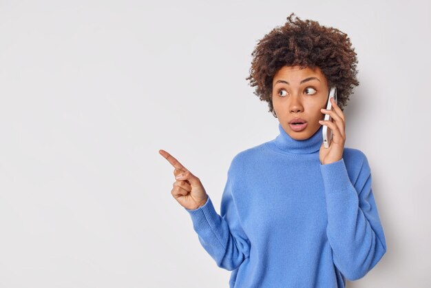 Beeindruckte schockierte junge Frau hat Telefongesprächspunkte Zeigefinger weg auf Leerzeichen hört schlechte Nachrichten, demonstriert Platz für Werbung einzeln auf weißem Hintergrund. Omg schau da