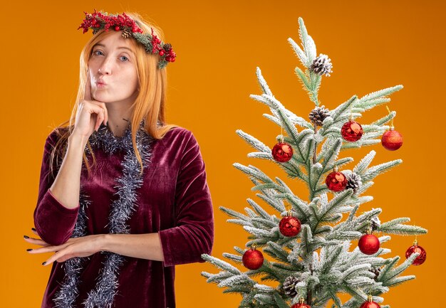 Beeindruckte spitzen Lippen junges schönes Mädchen, das nahe Weihnachtsbaum steht, das rotes Kleid und Kranz mit Girlande am Hals trägt Finger auf Wange lokalisiert auf orange Hintergrund