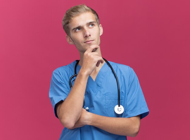 Beeindruckter Blick auf den jungen männlichen Arzt der Seite, der Arztuniform mit Stethoskop hält Kinn, das auf rosa Wand isoliert hält