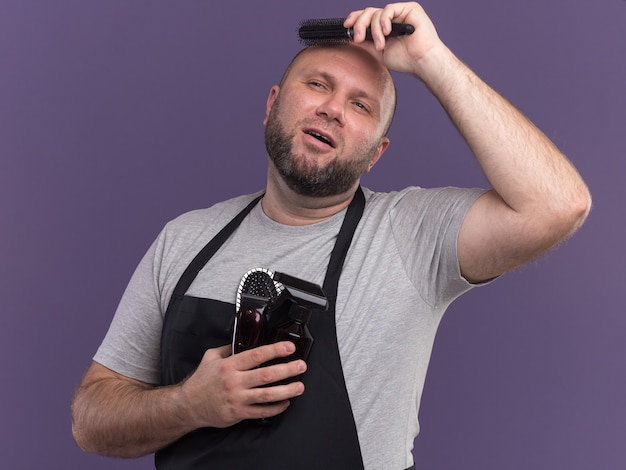 Beeindruckter Blick auf den seitlichen slawischen männlichen Friseur mittleren Alters in Uniform, der Friseurwerkzeuge hält und Haare kämmt, die auf lila Wand isoliert sind