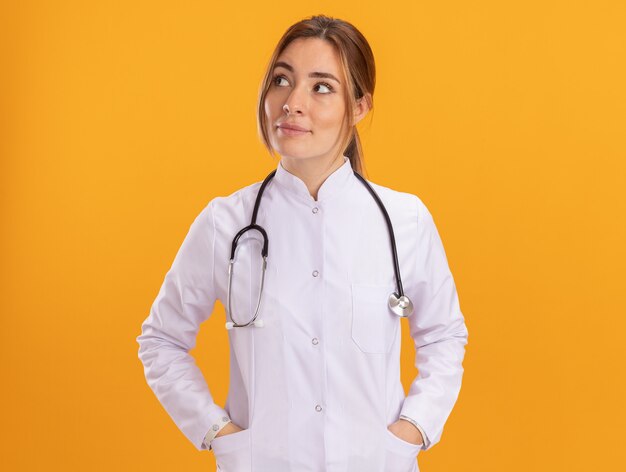 Beeindruckter Blick auf die junge Ärztin der Seite, die medizinische Robe mit Stethoskop trägt, die Hände auf Tasche lokalisiert auf gelber Wand setzt