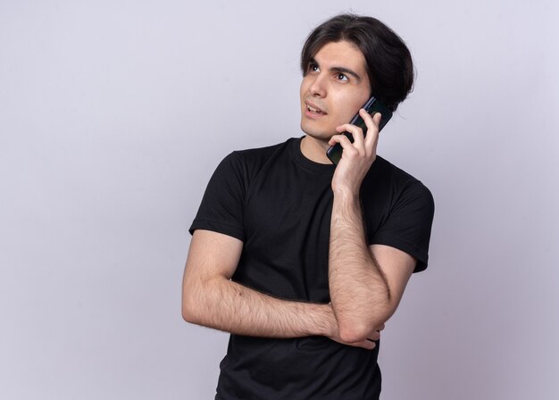 Beeindruckter Blick auf die Seite junger hübscher Kerl, der schwarzes T-Shirt trägt, spricht am Telefon lokalisiert auf weißer Wand