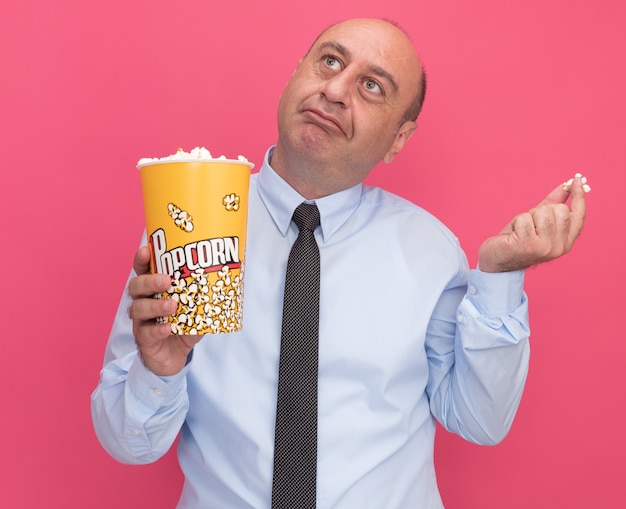 Beeindruckter Blick auf einen Mann mittleren Alters, der ein weißes T-Shirt mit Krawatte hält, das einen Eimer Popcorn mit einem Popcornstück hält, das auf rosa Wand lokalisiert wird
