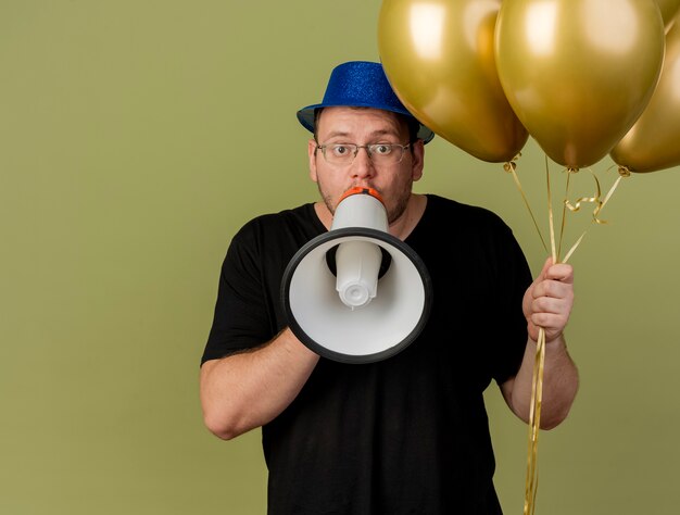 Kostenloses Foto beeindruckter erwachsener mann in optischer brille, die blauen partyhut trägt, hält heliumballons und spricht in lautsprecher, der auf olivgrüner wand isoliert wird