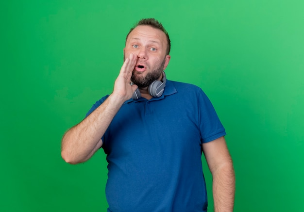 Beeindruckter erwachsener slawischer Mann, der Kopfhörer am Hals trägt und flüstert und Hand nahe Mund lokalisiert auf grüner Wand mit Kopienraum hält