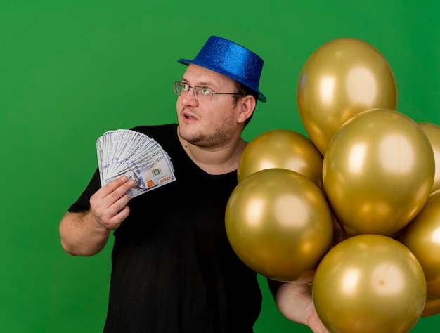 Kostenloses Foto beeindruckter erwachsener slawischer mann in optischer brille mit blauem partyhut hält heliumballons und geld auf der seite