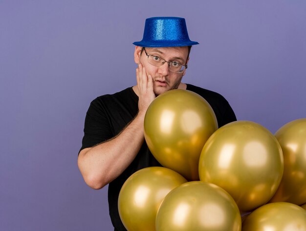 Kostenloses Foto beeindruckter erwachsener slawischer mann in optischer brille mit blauem partyhut legt hand auf gesicht und steht mit heliumballons balloon