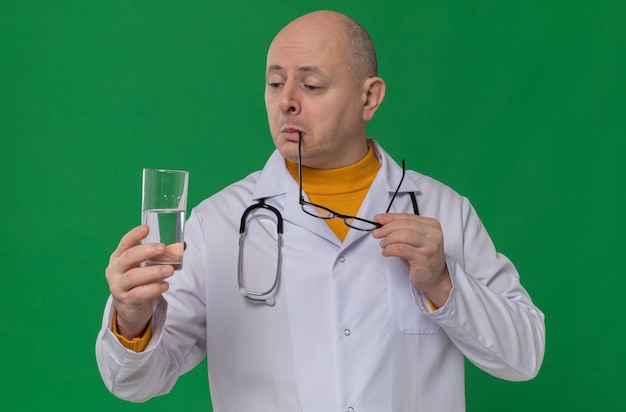 Beeindruckter erwachsener slawischer Mann mit optischer Brille in Arztuniform mit Stethoskop, das ein Glas Wasser hält und betrachtet