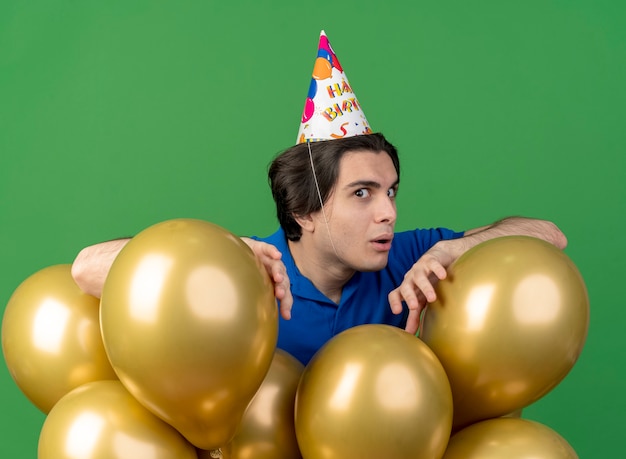 Beeindruckter gutaussehender kaukasischer Mann mit Geburtstagsmütze steht mit Heliumballons