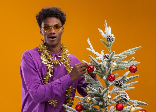 Beeindruckter junger afroamerikanischer Mann, der Brille mit Lametta-Girlande um den Hals trägt, der nahe verziertem Weihnachtsbaum auf orangefarbenem Hintergrund steht