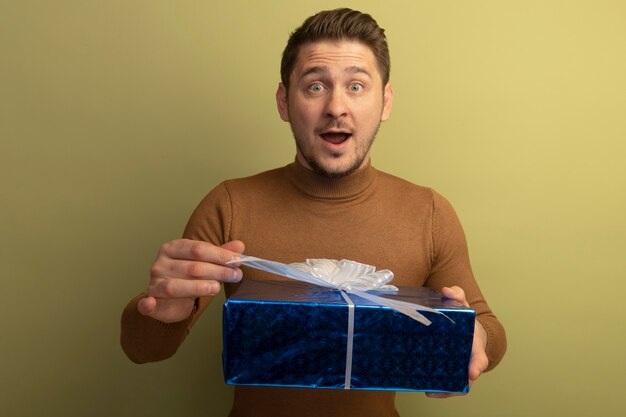 Beeindruckter junger blonder, gutaussehender Mann, der ein Geschenkpaket hält, das sein Band greift und nach vorne isoliert auf olivgrüner Wand schaut