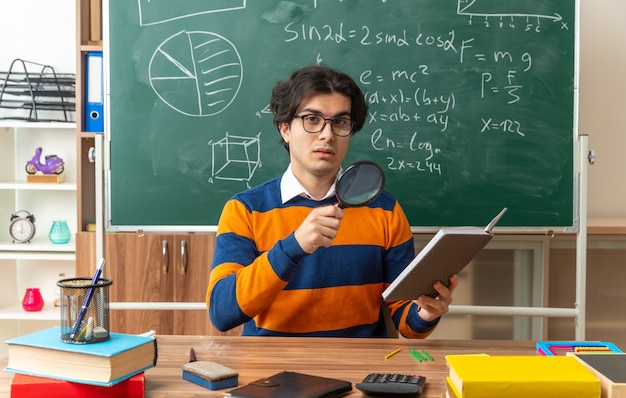 Beeindruckter junger Geometrielehrer mit Brille, der am Schreibtisch mit Schulmaterial im Klassenzimmer sitzt und Notizblock und Lupe mit Blick auf die Vorderseite hält