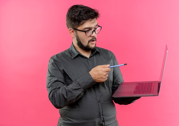 Beeindruckter junger Geschäftsmann, der Brillen hält und Punkte mit Stift am Laptop lokalisiert auf rosa Wand trägt