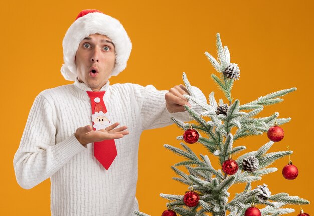 Beeindruckter junger gutaussehender Kerl mit Weihnachtsmütze und Weihnachtsmann-Krawatte, der in der Nähe des Weihnachtsbaums steht und ihn mit Weihnachtskugelverzierung verziert, die leere Hand isoliert auf oranger Wand zeigt