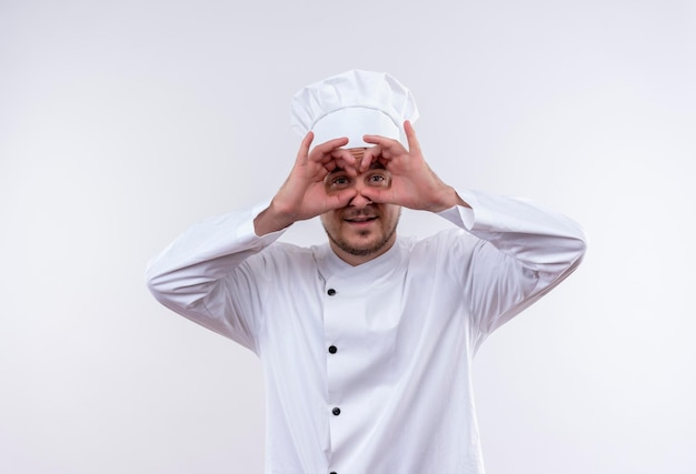 Beeindruckter junger gutaussehender Koch in Kochuniform, der die Blickgeste mit den Händen als Fernglas auf isolierter weißer Wand macht