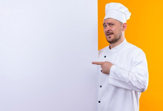Beeindruckter junger gutaussehender Koch in Kochuniform, der hinter weißer Wand steht und auf ihn isoliert auf oranger Wand mit Kopierraum zeigt