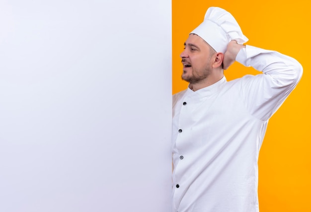 Beeindruckter junger gutaussehender koch in kochuniform, der hinter weißer wand steht und mit der hand darauf zeigt, isoliert auf oranger wand mit kopierraum
