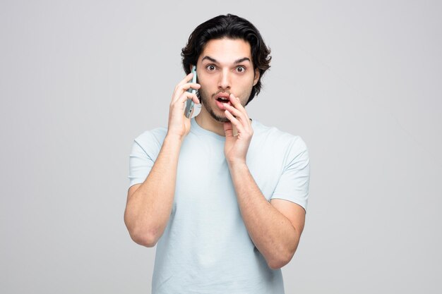 Beeindruckter junger gutaussehender Mann, der die Hand in der Nähe des Mundes hält und in die Kamera schaut, die am Telefon spricht, isoliert auf weißem Hintergrund