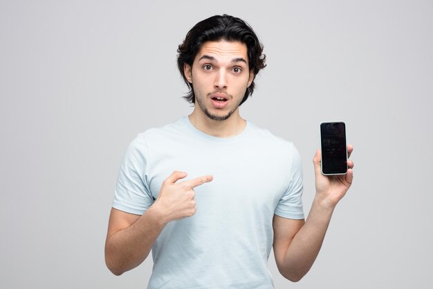 Beeindruckter junger gutaussehender Mann, der ein Handy zeigt, das darauf zeigt und die Kamera isoliert auf weißem Hintergrund betrachtet