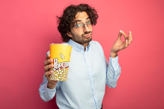 Beeindruckter junger gutaussehender Mann, der eine Brille trägt, die einen Eimer Popcorn und ein Popcornstück hält, das Front lokalisiert auf rosa Wand betrachtet
