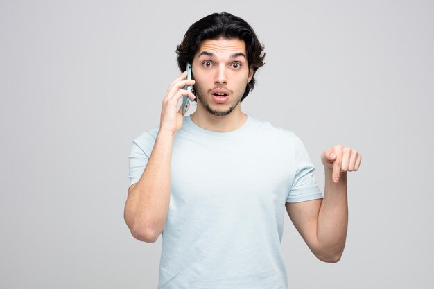 Beeindruckter junger gutaussehender Mann, der in die Kamera schaut, die nach unten zeigt, während er am Telefon spricht, isoliert auf weißem Hintergrund