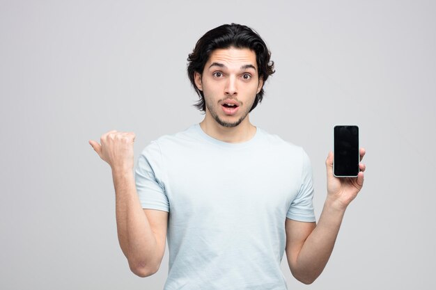 Beeindruckter junger gutaussehender Mann, der in die Kamera schaut und ein Handy zeigt, das auf die Seite zeigt, die auf weißem Hintergrund isoliert ist