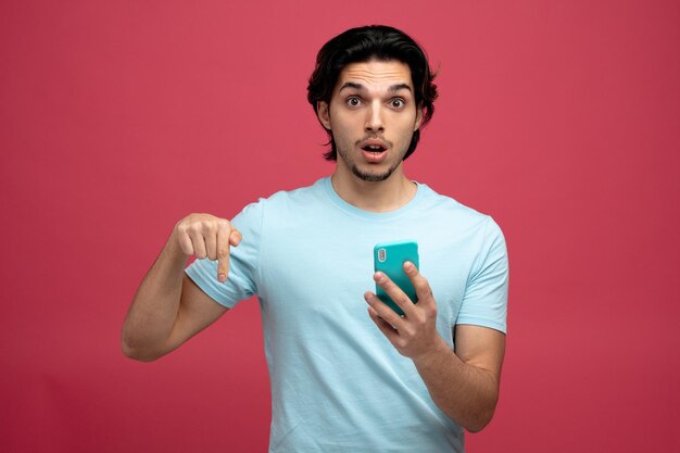 Beeindruckter junger gutaussehender Mann mit Handy, der auf die Kamera blickt, die nach unten zeigt, isoliert auf rotem Hintergrund