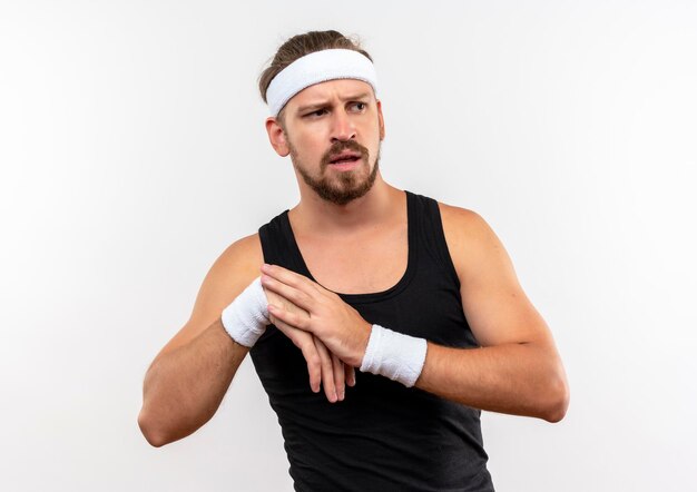 Beeindruckter junger, gutaussehender, sportlicher Mann mit Stirnband und Armbändern, der auf die Seite schaut und die Hand auf eine andere isoliert auf weißer Wand legt