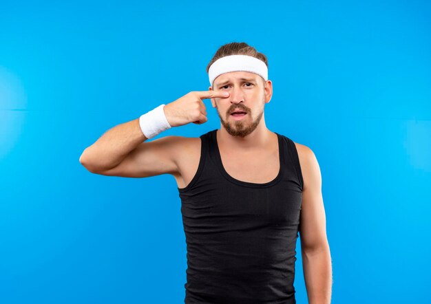 Beeindruckter junger, gutaussehender, sportlicher Mann mit Stirnband und Armbändern, der auf seine Nase zeigt, isoliert auf blauer Wand