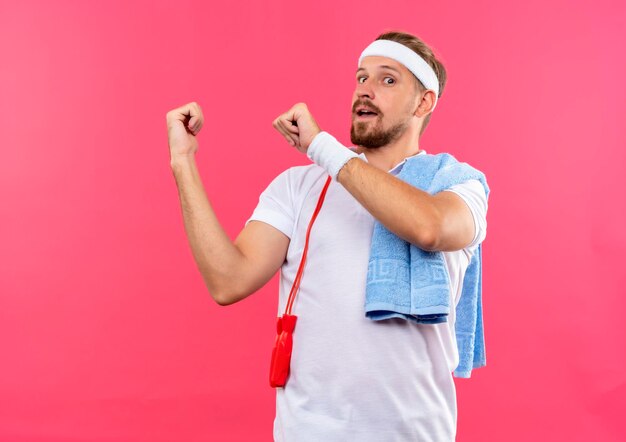 Beeindruckter junger, gutaussehender, sportlicher Mann mit Stirnband und Armbändern, der mit Springseil und Handtuch auf den Schultern isoliert auf rosa Wand zeigt