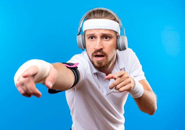 Beeindruckter junger, gutaussehender, sportlicher Mann mit Stirnband und Armbändern und Kopfhörern mit Armbinde, der auf die Seite isoliert auf blauer Wand zeigt