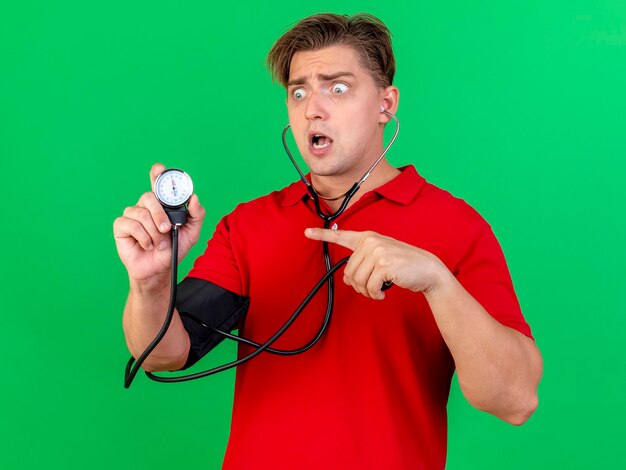 Beeindruckter junger hübscher blonder kranker Mann, der Stethoskop trägt, das Druck zu sich selbst hält, das Betrachten und Zeigen auf Blutdruckmessgerät lokalisiert auf grüner Wand