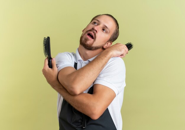 Beeindruckter junger hübscher Friseur, der Kämme hält, die lokal auf olivgrün mit Kopienraum suchen