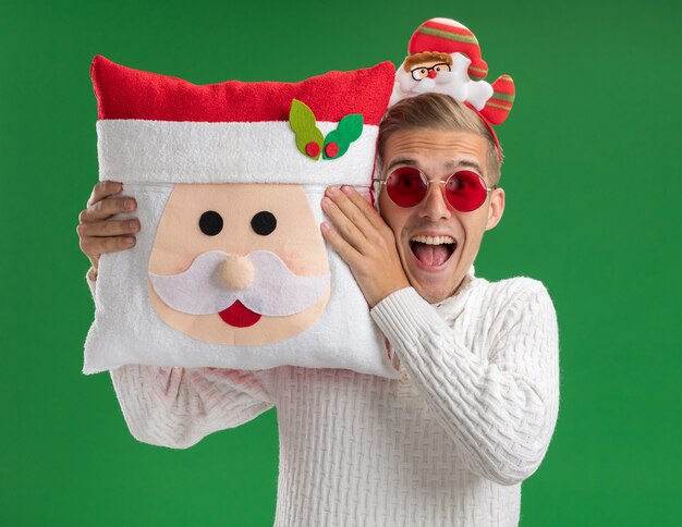 Beeindruckter junger hübscher Kerl, der Weihnachtsmann-Stirnband mit Brille trägt, die Weihnachtsmann-Kissen nahe Kopf lokalisiert auf grüner Wand hält