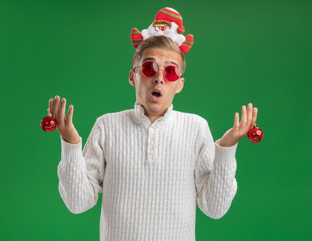 Beeindruckter junger hübscher Kerl, der Weihnachtsmann-Stirnband mit Brille trägt, die Weihnachtsverzierungskugeln hält, die Kamera lokalisiert auf grünem Hintergrund betrachten