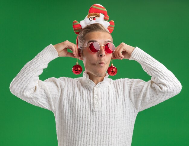Beeindruckter junger hübscher Kerl, der Weihnachtsmann-Stirnband mit Gläsern hängenden Weihnachtskugeln auf Ohren trägt, die Seite mit gespitzten Lippen lokalisiert auf grünem Hintergrund betrachten