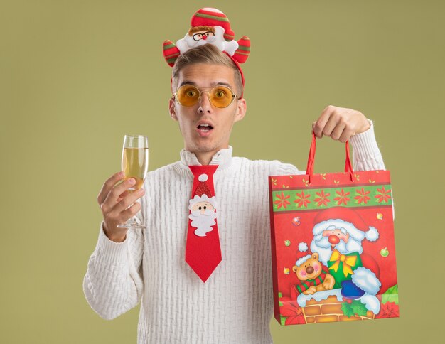 Beeindruckter junger hübscher Kerl, der Weihnachtsmann-Stirnband und Krawatte hält, die Glas Champagner und Weihnachtsgeschenkbeutel lokalisiert auf olivgrüner Wand hält