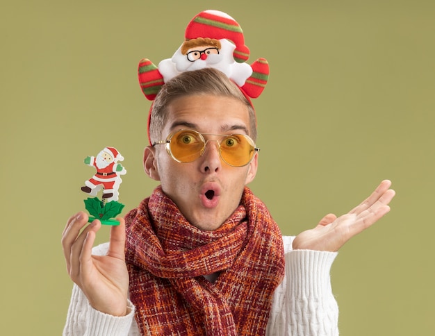 Beeindruckter junger hübscher Kerl, der Weihnachtsmann-Stirnband und Schal hält, der Weihnachtsmann-Spielzeug hält, das leere Hand lokalisiert auf olivgrüner Wand zeigt