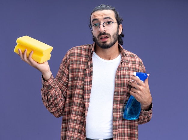 Kostenloses Foto beeindruckter junger hübscher putzmann, der t-shirt hält, das sprühflasche mit schwamm lokalisiert auf blauer wand hält