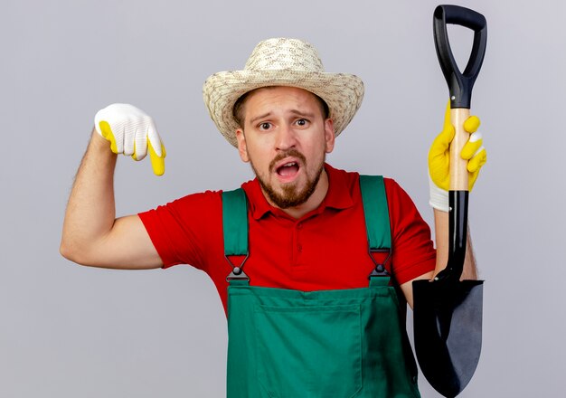 Beeindruckter junger hübscher slawischer Gärtner in der Uniform, die Gartenhandschuhe und Hut hält, der Spaten hält, der unten auf weißer Wand zeigt