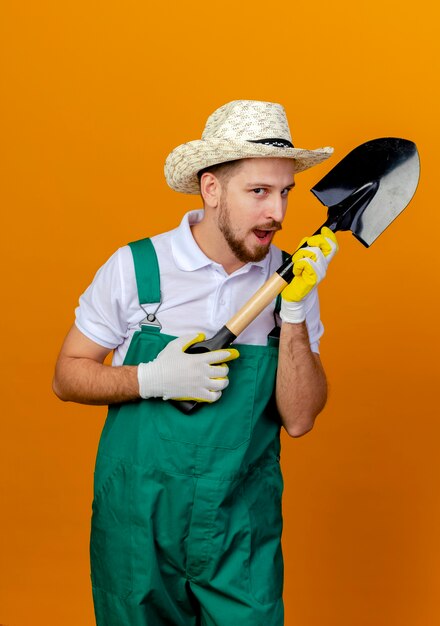Beeindruckter junger hübscher slawischer Gärtner in der Uniform, die Hut und Gartenhandschuhe trägt, die Spaten halten, der lokal auf orange Wand schaut